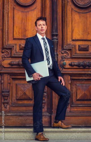 American Businessman working in New York, wearing black suit, patterned necktie, white undershirt, brow leather shoes, holding laptop computer, standing on stairs by office doorway, looking forward