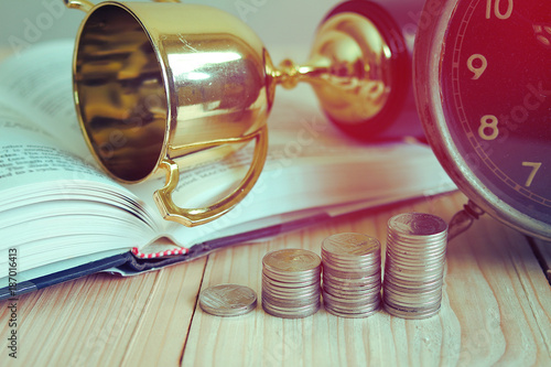 Stack of coin on the wood table with background clock and trophy lay on book , finance concept. photo