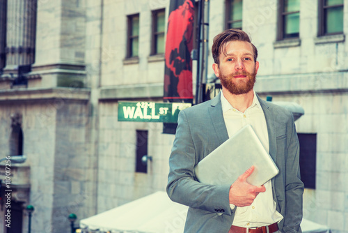 American Businessman with beard, mustache traveling, working in New York, wearing cadet blue suit, white undershirt, carrying laptop computer, standing on Wall Street by street sign, looking forward. photo