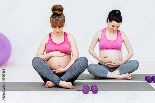 Sport, therapy and fitness concept. Two young pregnant women doing relaxation exercise on exercising mat on white background.