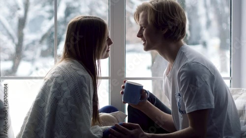 Great snowfall outside. Two belovers spend time in the warm room at the window with smelling coffee cups.