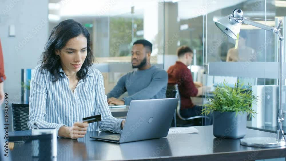 Beautiful Female Office Worker Makes Internet Purchase from Her Laptop Using Credit Card. In the Background Creative Young People Working. Shot on RED EPIC-W 8K Helium Cinema Camera. Beautiful Female Office Worker Makes Internet Purchase from Her Laptop Using Credit Card. In the Background Creative Young People Working. Shot on RED EPIC-W 8K Helium Cinema Camera.