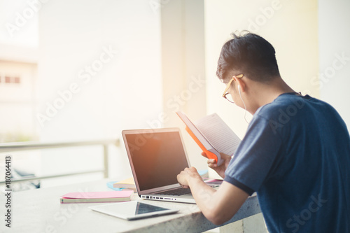 College student boy reading book for exam and listening to music via headphones with laptop.