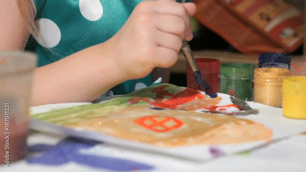 Close-up of hands of unidentified little child dressed in polka dots dress sitting at table in kindergarten and drawing picture of house and family with brush and watercolours. Close-up of hands of unidentified little child dressed in polka dots dress sitting at table in kindergarten and drawing picture of house and family with brush and watercolours.