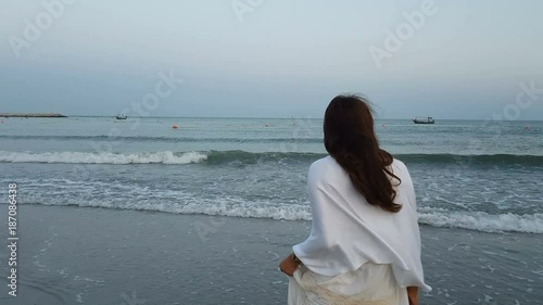 back view of young beautiful asian female standing on the beach and wind blow her hair while she is looking to the seascape and skyscape in the evening at dusk. relaxation, lifestyle, travel concept.