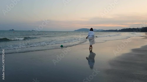 back view of young asian beautiful female in white dress walking barefoot on wet sandy beach while tide or wave is crashing to the her foots and beach when twilight or dusk.