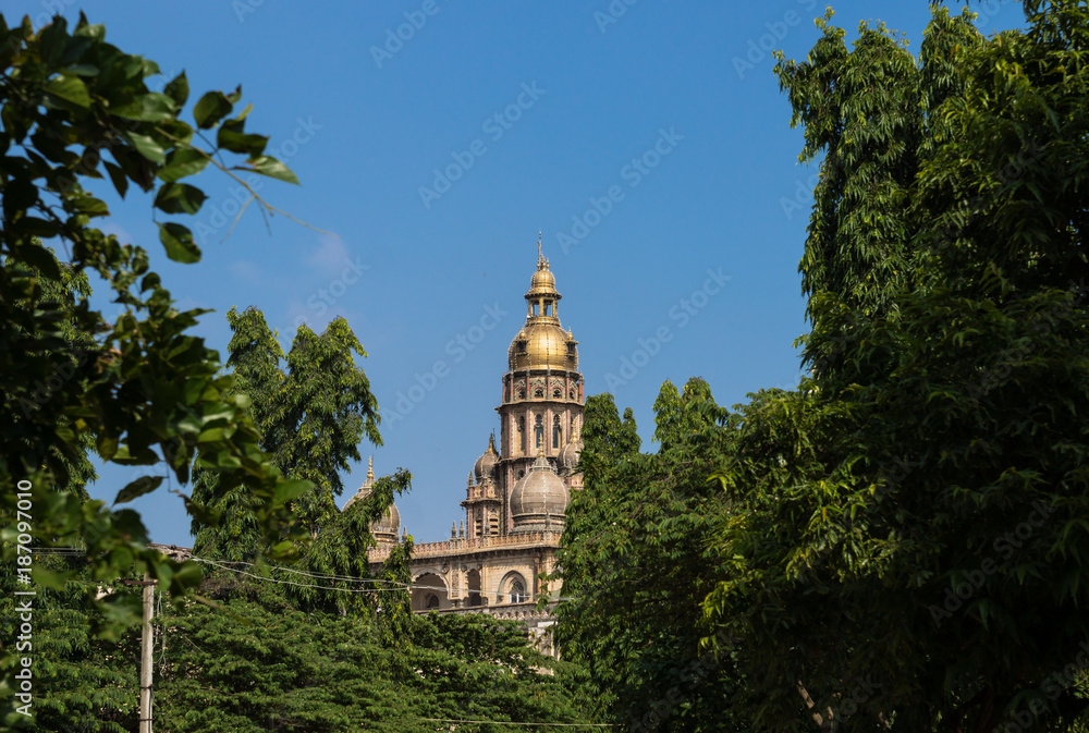 Golden tower of Mysore Palace behind green trees with blue sky, Mysore, India