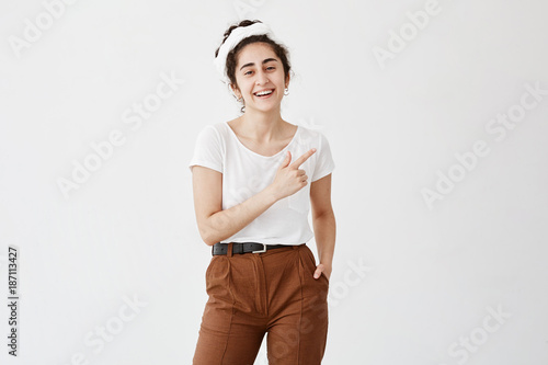 Advertising concept. Indoor shot of positive friendly young female with dark wavy hair in bun pointing at copy space on white background for your text or promotional content