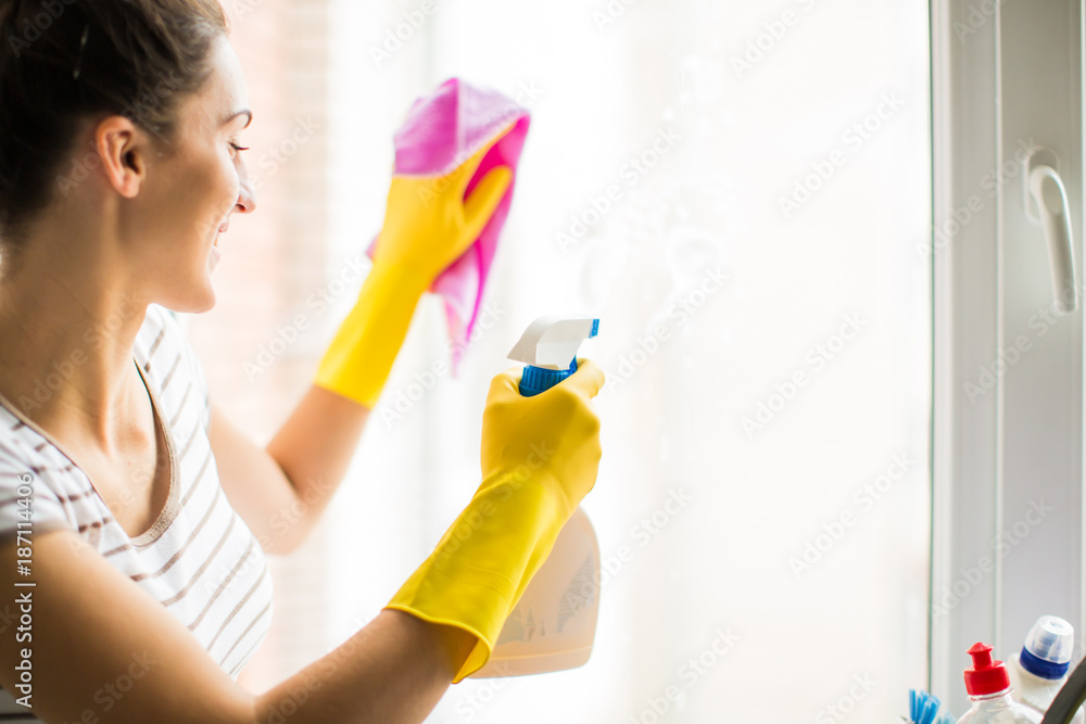 The young smiling woman in gloves washing the window at home The young smiling woman in gloves washing the window at home