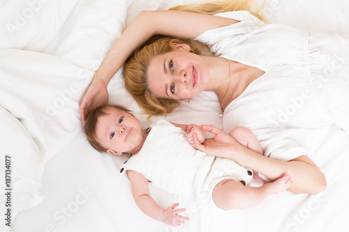Portrait of young mother with blond hair with her sweet 3 month old baby girl in white wear having fun in the bedroom at morning, loving happy family concept, top view