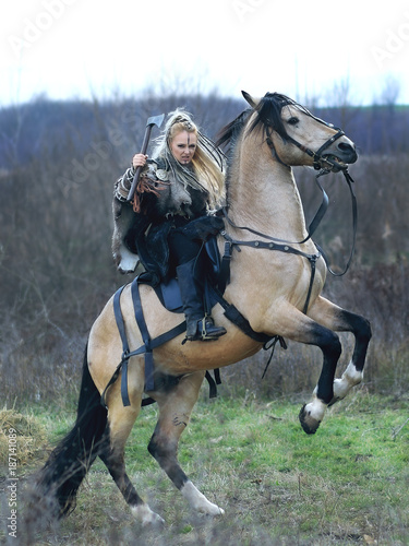 Beautiful viking warrior woman with ax in traditional warrior clothes riding a horse, threatening and ready to battle