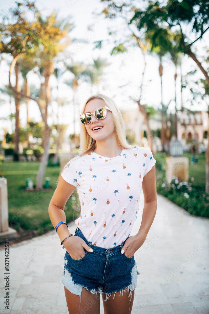 Fototapeta premium Attractive blonde woman in stylish sunglasses posing outside with exotic palm trees on background. Portrait of young woman with cute hairstyle walking down the street