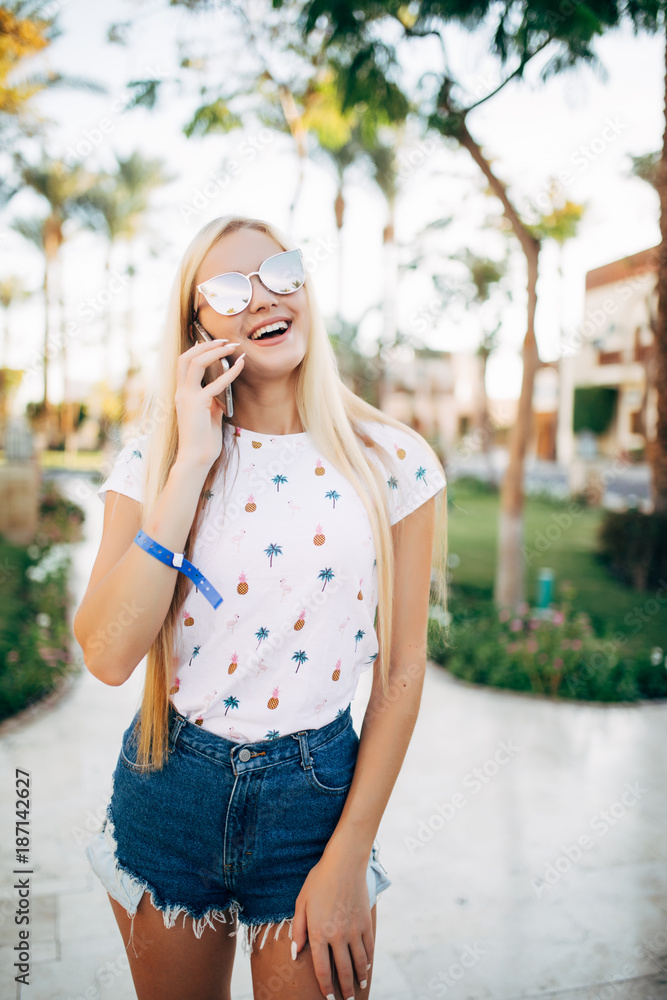 Portrait of young style woman on the road with palm trees. She talks on the phone with charming smile and looks happy. Sunny summer day. Vacation mood.
