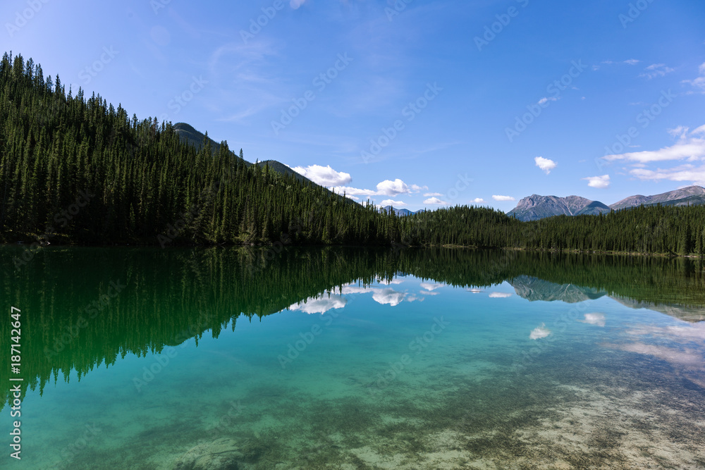A turquoise alpine lake reflects the treeline perfectly. The sky is almost clear.