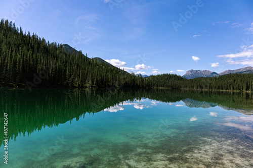 A turquoise alpine lake reflects the treeline perfectly. The sky is almost clear. © Reid