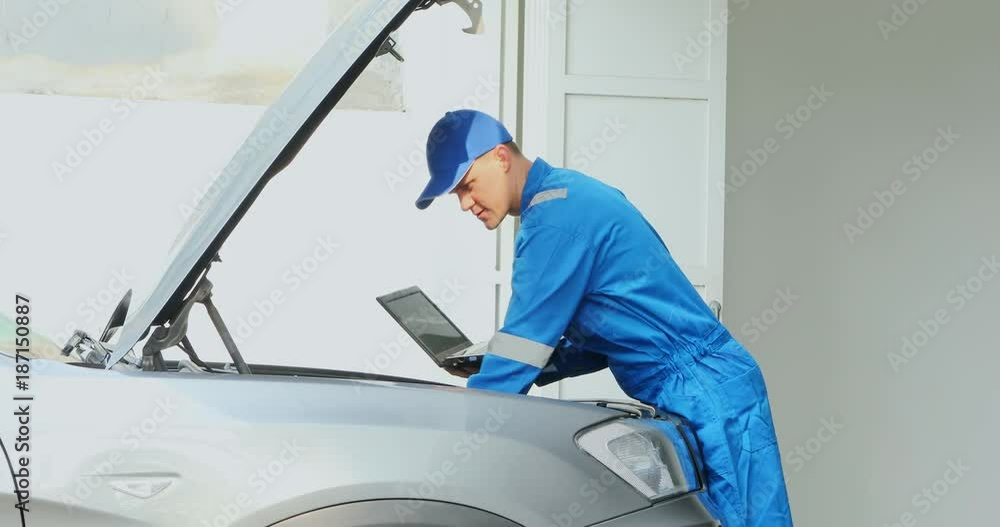 Male Caucasian mechanic checking a broken car machine while holding a laptop computer in the garage Male Caucasian mechanic checking a broken car machine while holding a laptop computer in the garage