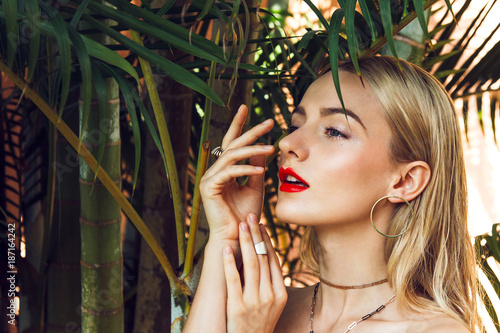 Close-up portrait of a beautiful young woman with long blonde hair, green mysterious eyes and red big sexy lips posing next to the palms tree. Her looks is complemented by golden accessories.