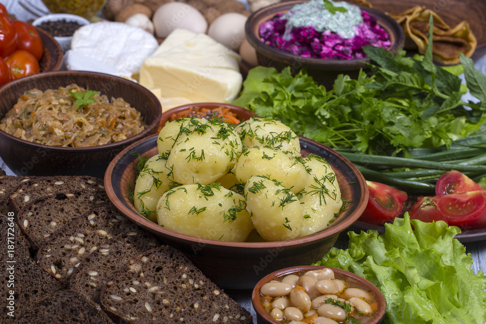 Group food on table: boiled young potatoes, beans, braised cabbage, beet salad with cottage cheese, sliced cheeses, tomato, korean carrot, black bread with seeds, green lettuce leaves, spice, egg