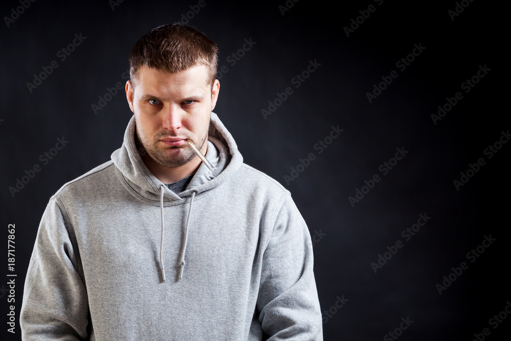 A young, dark-haired, sad-looking man in a sporty gray sweatshirt fell ill with a cold, measured the temperature on a black isolated background. A man holding a thermometer in his mouth