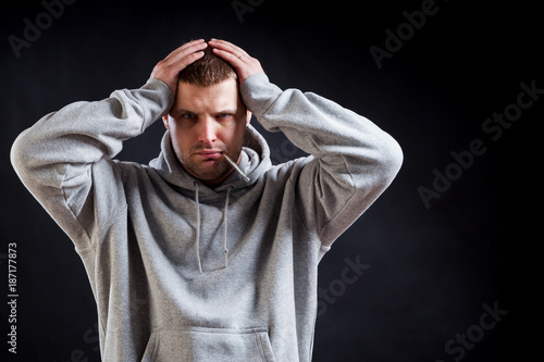 A young, dark-haired, sad man in a sporting gray sweatshirt fell ill with a cold, his head hurts and he holds onto it on a black isolated background. The man measures the temperature © Виталий Сова