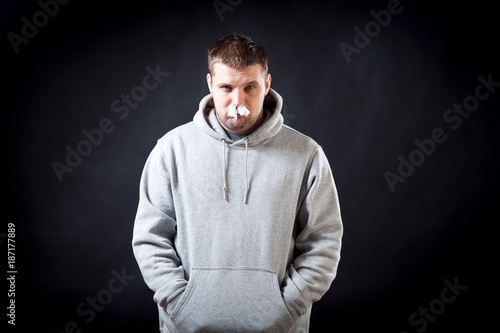 A young dark-haired man in a sporting gray sweatshirt fell ill with a cold, he had a nose on a black isolated background. The man in the nose stuffed napkins, so as not to flow snot