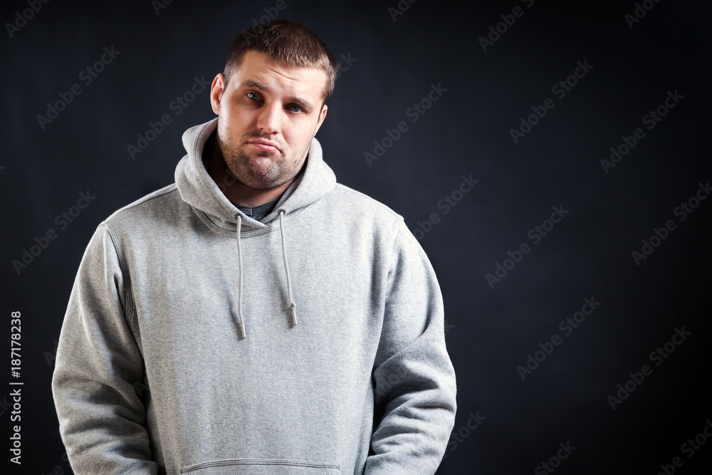 A young dark-haired man in a sports gray sweatshirt was upset that he fell ill and looks sad, his hands are holding in the pockets on a black isolated background A young dark-haired man in a sports gray sweatshirt was upset that he fell ill and looks sad, his hands are holding in the pockets on a black isolated background
