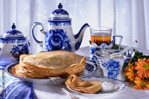 Freshly baked pancakes, a teapot, a glass with tea, sour cream are on a table set for the maslenitsa, pancake week celebration