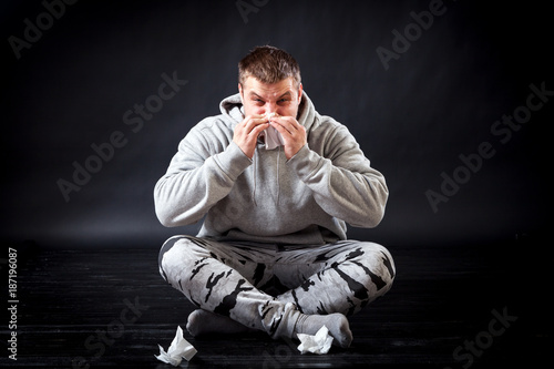 A young sad man in sports clothes sits on the floor and is upset that he is sick, his snot is flowing, he is holding a napkin and blowing his nose in a black isolated background