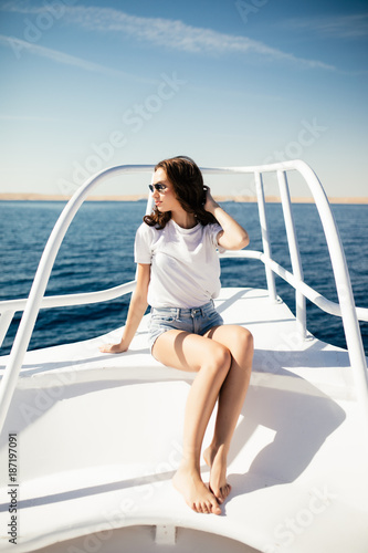 Young woman in summer outfit sitting at deck of yacht and looking away with sea on background