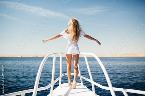 Woman standing on the nose of the yacht at a sunny summer day, breeze developing hair with open arms beautiful sea on background