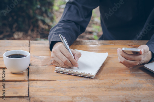 Fotografie Close up businessman write notebook and using cellphone on wood table
