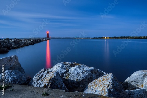 Lighthouse Beacon At Night. The guiding red light of the Manistique Lighthouse reflects on the calm blue water of a scenic Great Lakes Harbor in the Upper Peninsula of Michigan.