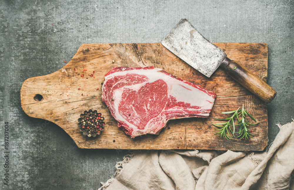 Fototapeta premium Flat-lay of raw prime beef meat dry-aged steak rib-eye with seasoning and chopper knife on wooden cutting board over grey concrete background, top view. Meat high-protein dinner concept