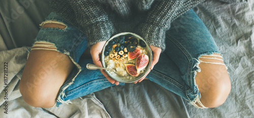 Healthy winter breakfast in bed. Woman in sweater and jeans holding rice coconut porridge with figs, berries, hazelnuts, top view, wide composition. Clean eating, vegetarian, comfort food concept
