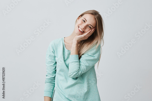 Excited overjoyed beautiful blonde woman keeps hand on cheek, smiles with enjoyment as notices something pleasant, isolated against gray background with copy space for promotional text