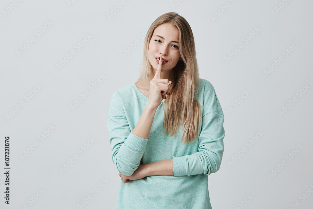 Horizontal shot of pretty beautiful female keeps finger on smiling lips, demonstrates hush sign, asks to keep conspiracy or silence, tries to hide secret, isolated against gray studio background Horizontal shot of pretty beautiful female keeps finger on smiling lips, demonstrates hush sign, asks to keep conspiracy or silence, tries to hide secret, isolated against gray studio background