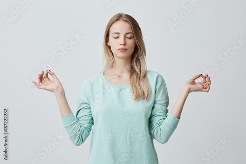 Restful peaceful blonde female feels relaxed, stands in lotus pose, tries to concentrate or to be focused, closes eyes, enjoys silence, tries to find balance. Calm atmosphere and meditation photo