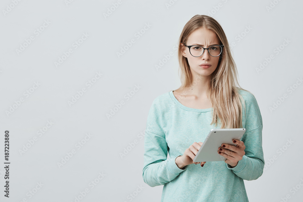 Attractive blonde office worker in stylsh eyewear works, look at camera with appeal, holds tablet in hands, surfs internet. Tired Caucasian woman dressed in blue sweater uses modern technologies. Attractive blonde office worker in stylsh eyewear works, look at camera with appeal, holds tablet in hands, surfs internet. Tired Caucasian woman dressed in blue sweater uses modern technologies.