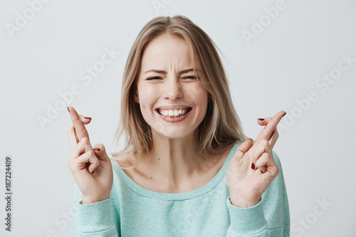 Young caucasian blonde female closing her eyes crossing fingers with hope, anticipating important news. Beautiful girl with hopeful expression gesturing with fingers, showing white teeth