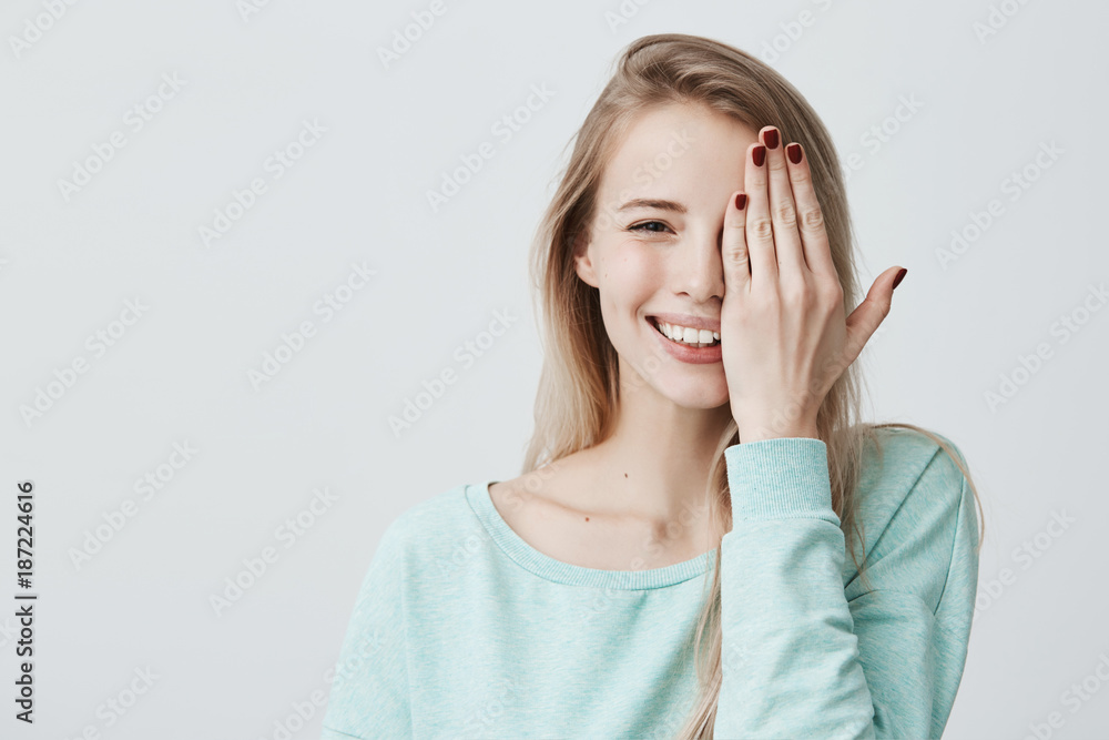 Glad caucasian woman with long dyed hair, wearing light blue sweater, isolated against gray background, closing her eye with hand. Happy positive female having good and playful mood. Glad caucasian woman with long dyed hair, wearing light blue sweater, isolated against gray background, closing her eye with hand. Happy positive female having good and playful mood.