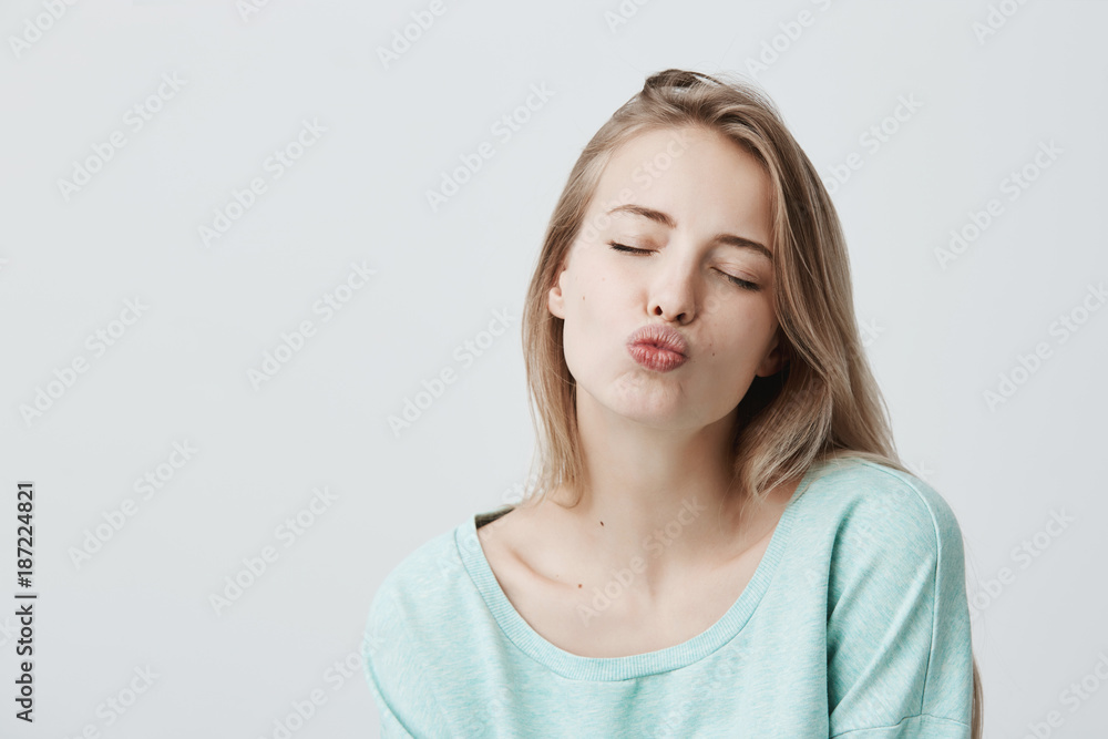 Beautiful dreamy female with blonde hair closes eyes, pouts lips, sends kisses to camera, poses against gray background. Attractive charming woman with appealing appearance in light blue sweater Beautiful dreamy female with blonde hair closes eyes, pouts lips, sends kisses to camera, poses against gray background. Attractive charming woman with appealing appearance in light blue sweater