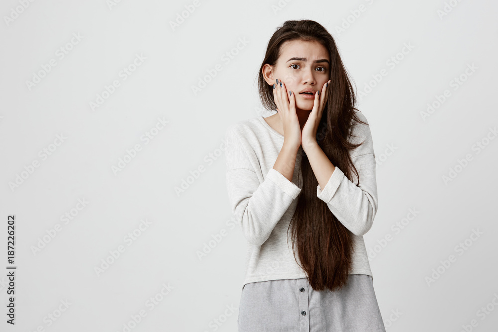 Brunette teenage girl with dark eyes raising eyebrows, staring with big eyes, holding hands on her cheeks, being in panic worried to hear bad news. Horrified young female with shocked scared look Brunette teenage girl with dark eyes raising eyebrows, staring with big eyes, holding hands on her cheeks, being in panic worried to hear bad news. Horrified young female with shocked scared look
