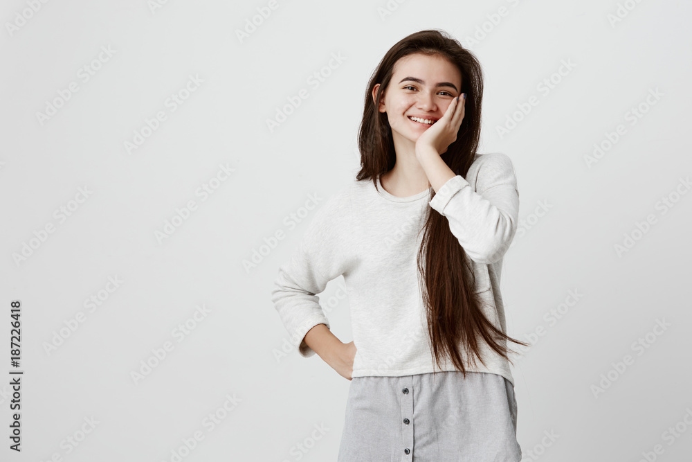 Waist-up portrait of brunette female model joyfully smiling with white teeth, glad to receive good news or present. Excited girl expresses positive emotinos, holds hand on cheek in amazement Waist-up portrait of brunette female model joyfully smiling with white teeth, glad to receive good news or present. Excited girl expresses positive emotinos, holds hand on cheek in amazement
