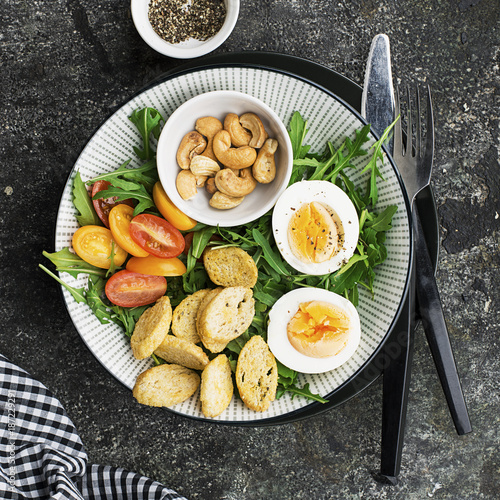 Fresh spring salad with boiled egg, arugula, salad, tomatoes, pecans and crispy toasted bread croutons in a large dish on a gray background. Top view..