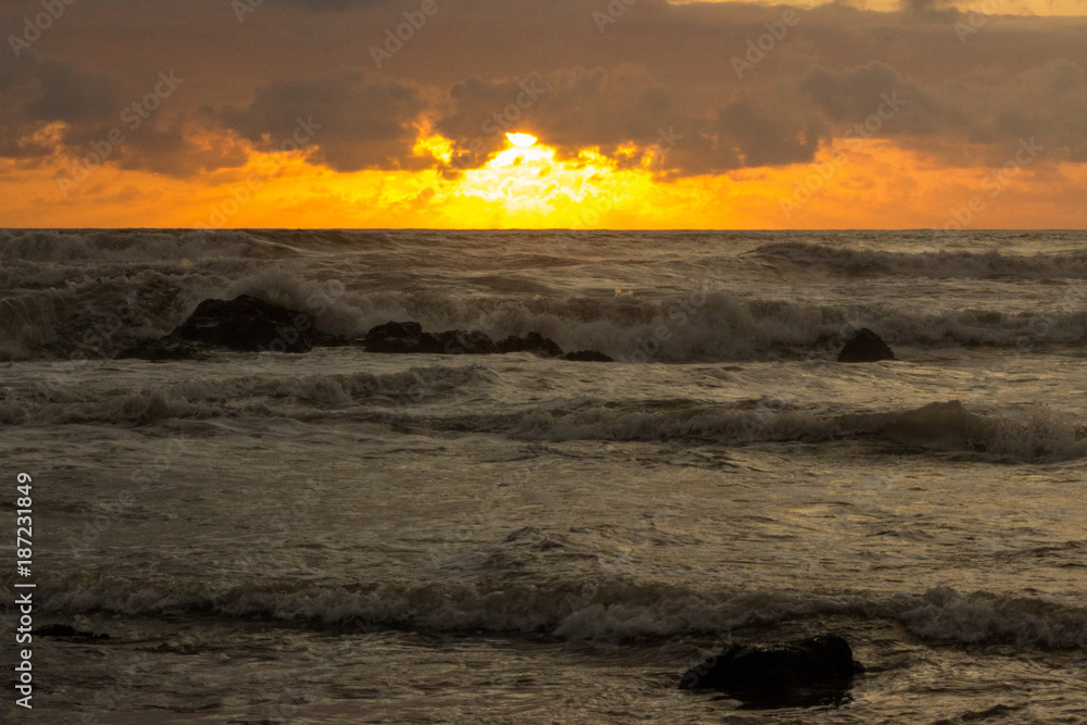Waves crash ashore as the Southern Pacific sun sets in the background, turning the sky to liquid orange fire that is reflected in the water.