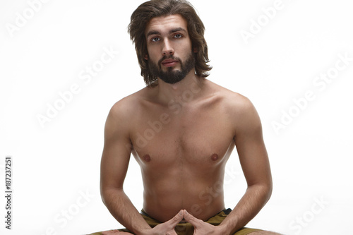 Waist up portrait of attractive young unshaven European male with thick beard and wavy hair posing shirtless in white studio. Bearded guy with naked athletic torso doing yoga in the morning