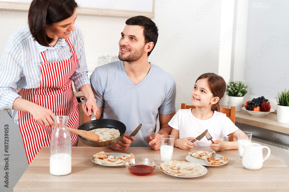 Photo of hard working wife, husband and their daughter sit together at kitchen table, going to eat delicious pancakes, enjoy togetherness. Friendly family eat breakfast, appetizing flannel cake. Photo of hard working wife, husband and their daughter sit together at kitchen table, going to eat delicious pancakes, enjoy togetherness. Friendly family eat breakfast, appetizing flannel cake.