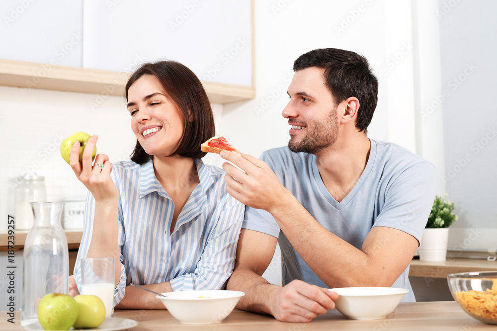 Happy couple spend free time or weekend together at kitchen, glad husband suggests wife to eat snack, she refuses as eats green apple, have breakfast. Family, cooking and relations concept.