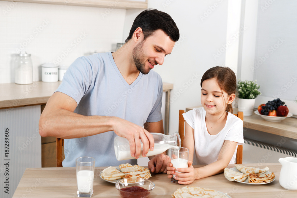 Happy father and daughter have breakfast at kitchen, eats delicious pancakes with jam, drink milk, enjoys delicious food prepared by mother. Family apetizes yummy sweet dish. Eating and people concept