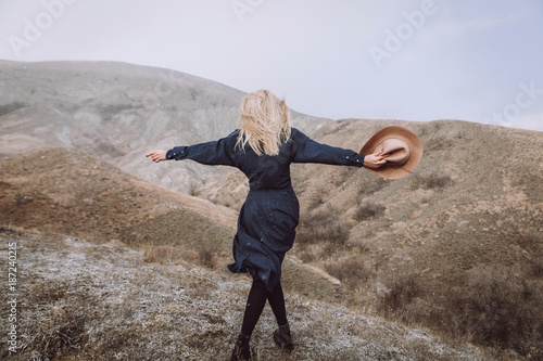Plakat stylowa blond dziewczyna w niebieskiej sukience i beżowym kapeluszu w dłoniach stoi z plecami w górach na łonie natury z pięknym widokiem i skandynawskim krajobrazem
